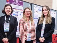 Three female student poster session participants standing in front of two posters